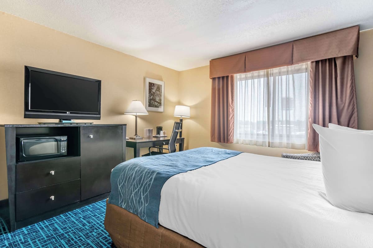 A hotel room with a bed, a desk with a computer, a television, a microwave, and a window with curtains.