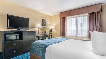 A hotel room with a bed, a desk with a computer, a television, a microwave, and a window with curtains.