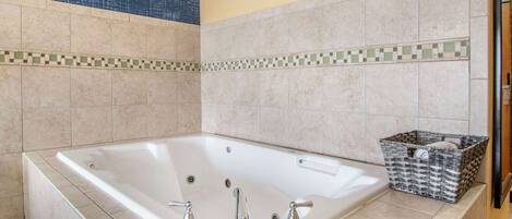 Una bañera grande y rectangular con dos grifos, una bandejita pequeña y una cesta de mimbre al lado.