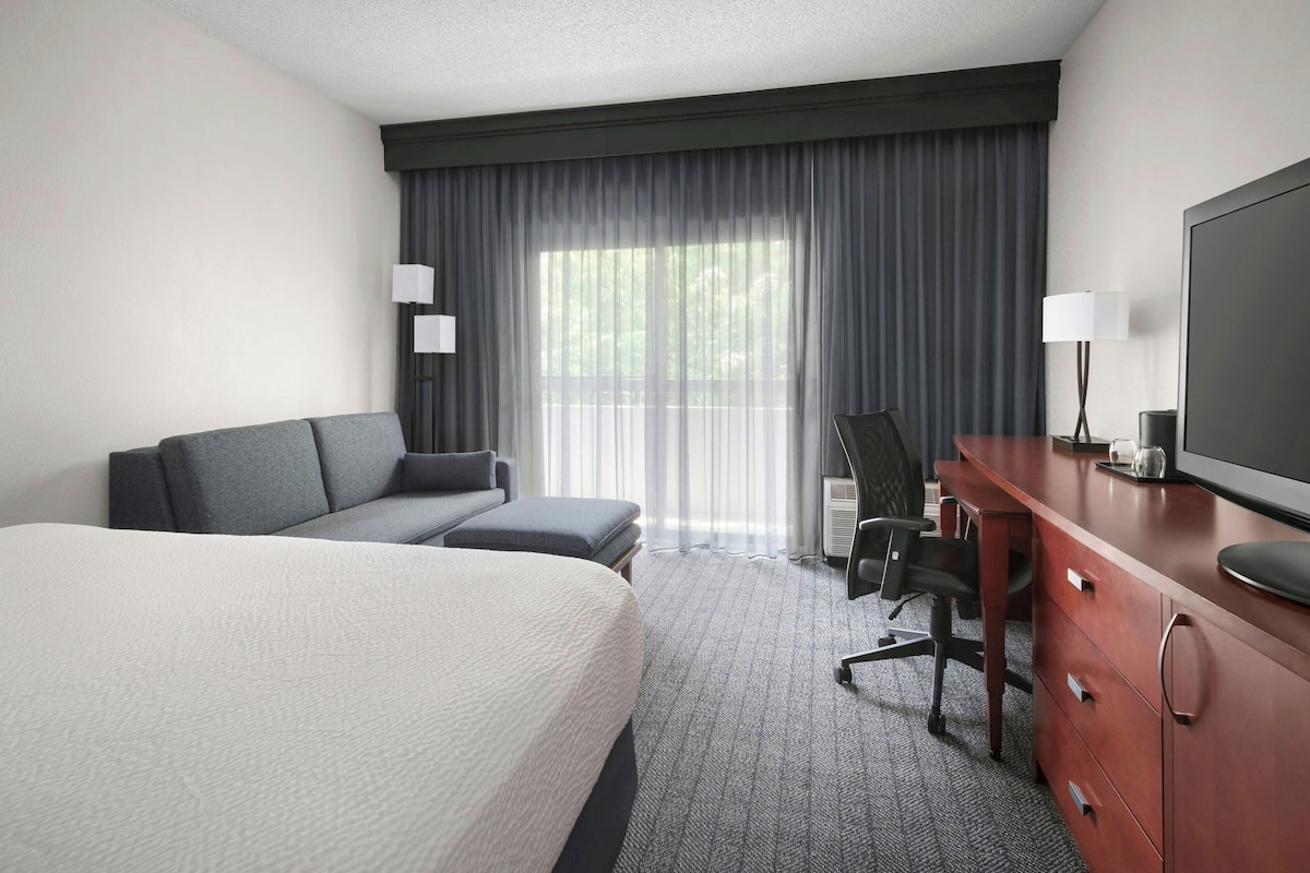 A hotel room with a large bed, a sofa, a desk with a chair, a television, and a window with curtains.