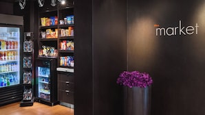 Un espacio comercial moderno con estantes llenos de refrigerios y bebidas, un expositor refrigerado y un jarrón decorativo con flores moradas.