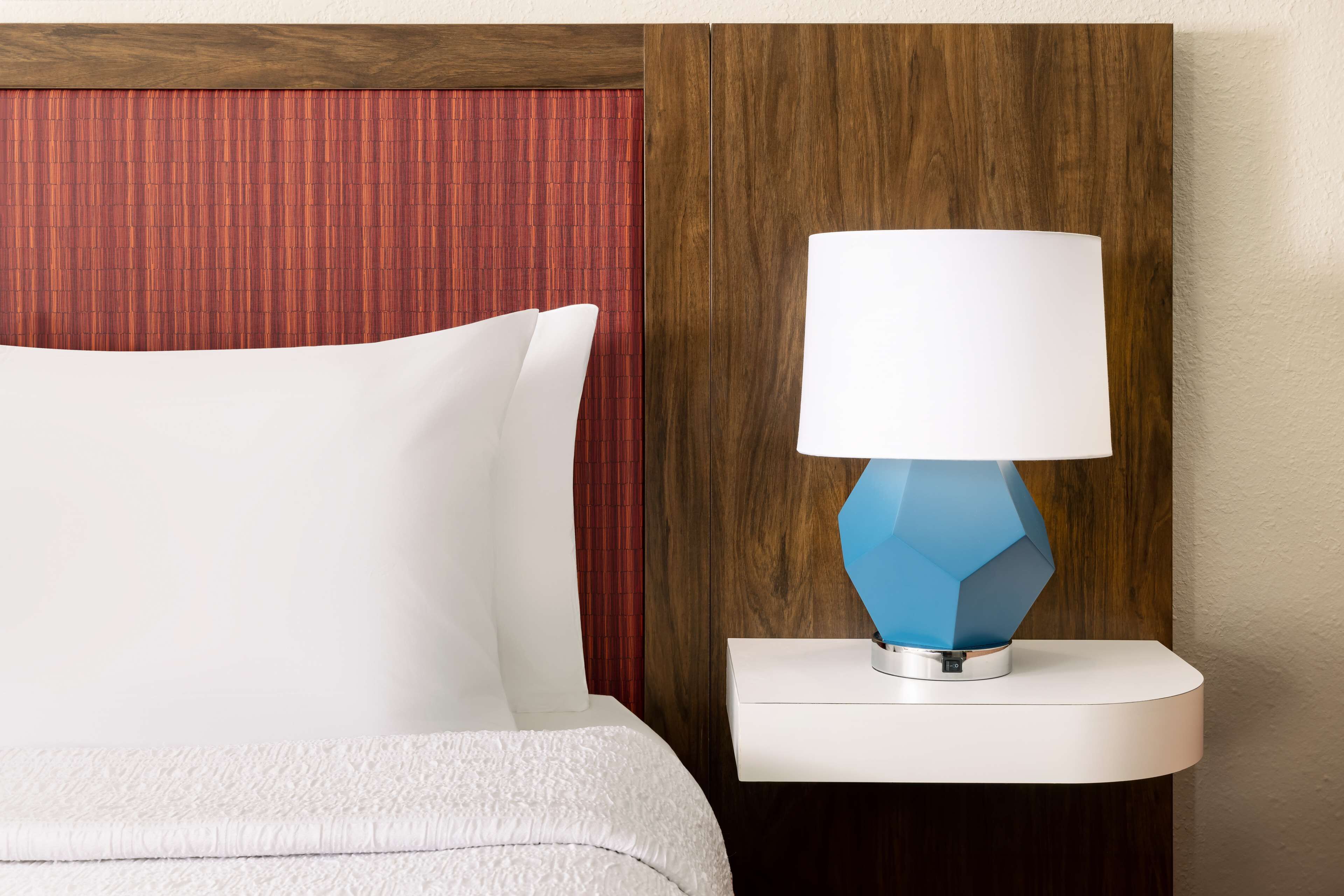 A bed with white bedding, a blue geometric lamp on a white bedside table, and a wooden headboard with a red panel.