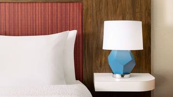 Una cama con ropa de cama blanca, una lámpara geométrica azul sobre una mesita de noche blanca, y un cabecero de madera con un panel rojo.