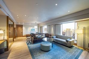 Una sala de estar moderna con sofá, reposapiés y mesa de centro, suelo de madera y ventanas grandes con vistas a la ciudad.