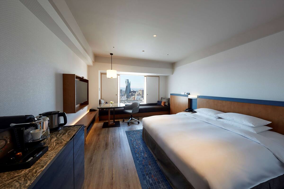 A hotel room with a bed, a desk with a coffee maker, a TV, and a view of the city.