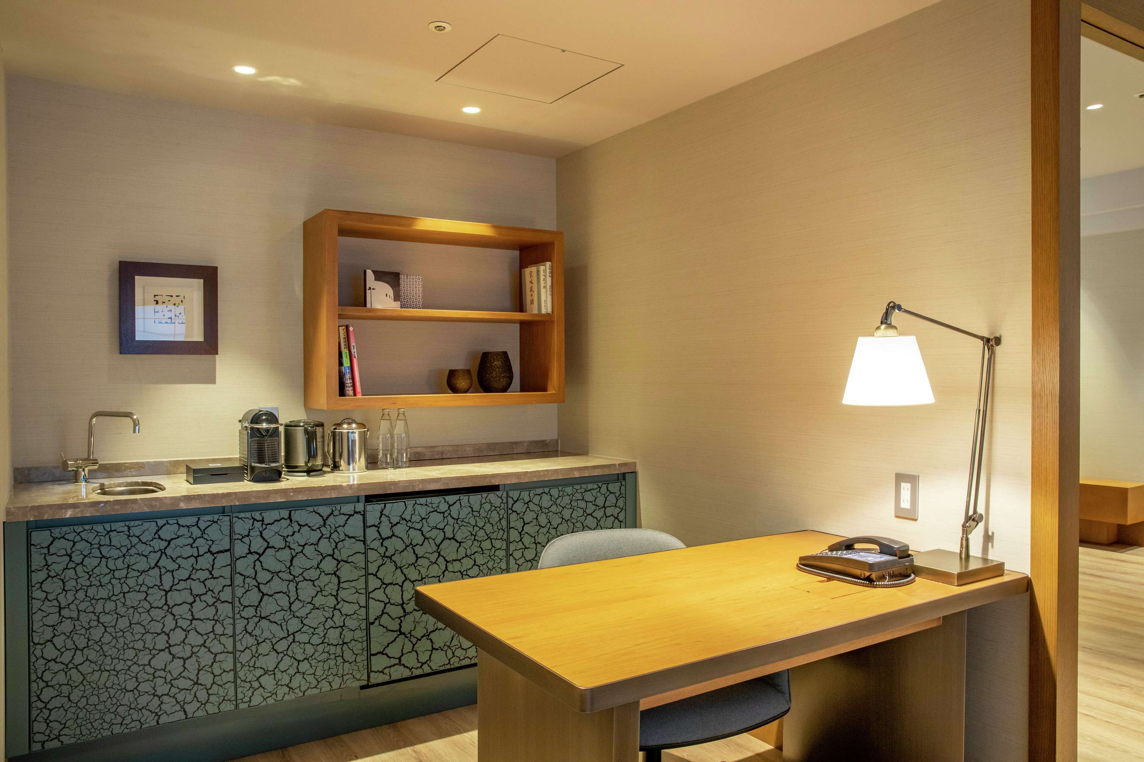 A modern kitchen with a wooden countertop, a built-in cabinet with a cracked tile backsplash, a small shelf with books, and a table with a lamp.