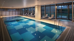Una amplia piscina interior con baldosas azules cristalinas, sillones de descanso y grandes ventanales con vistas al exterior.