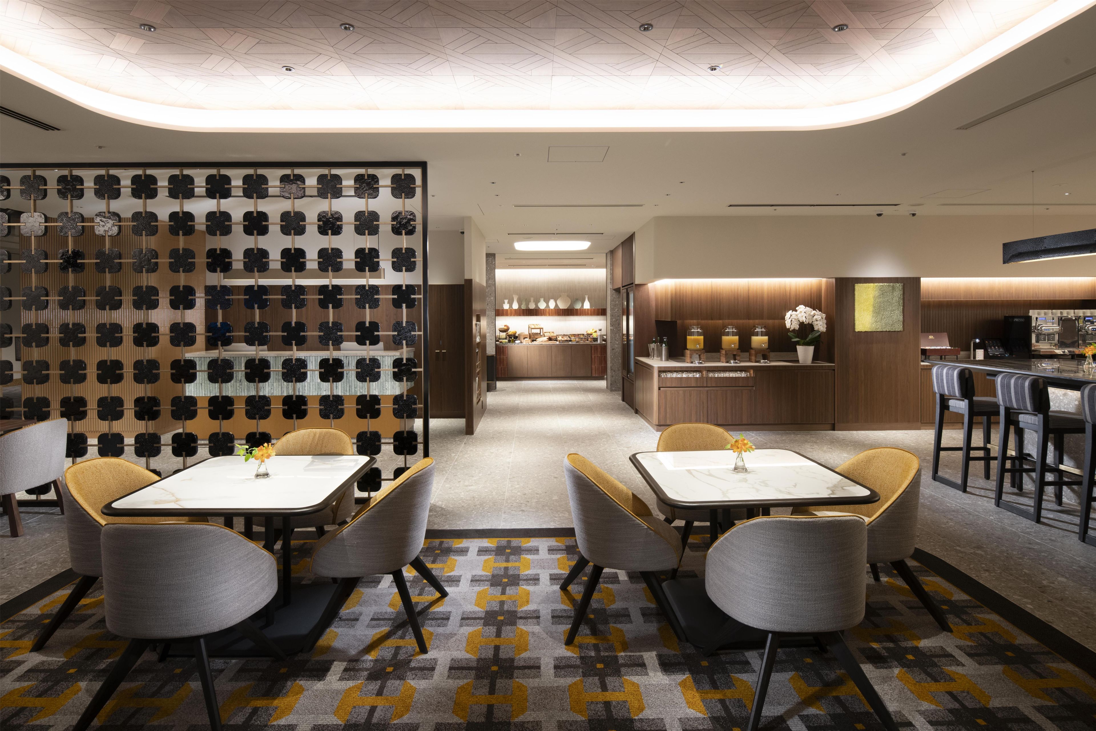 A modern hotel lobby with a central seating area, a bar, and a kitchen visible in the background.
