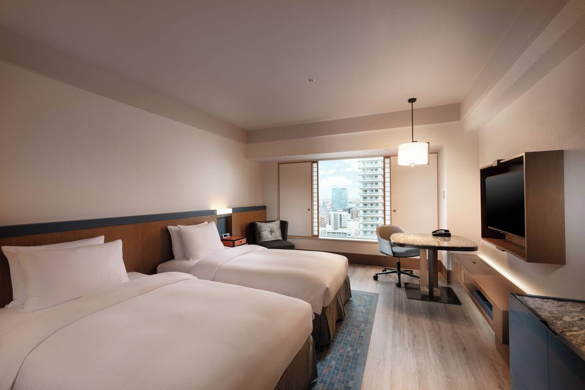 A hotel room with two beds, a desk, a chair, a TV, and a window with a city view.