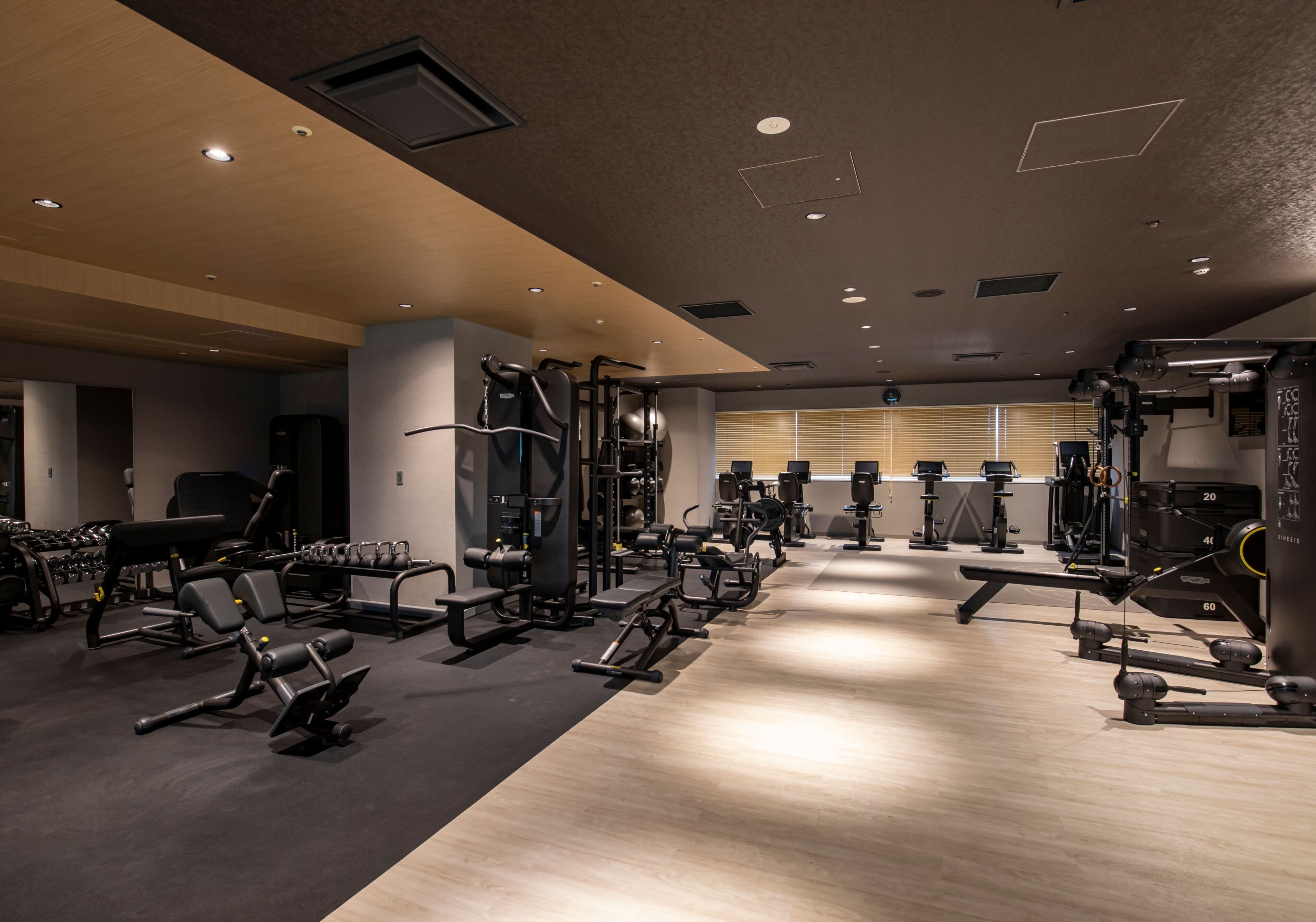 A modern gym with various exercise equipment, a wooden floor, and a large window with blinds.