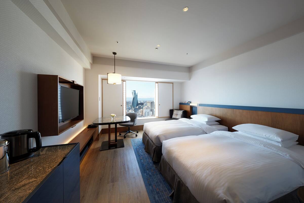 A hotel room with two beds, a TV, a desk with a chair, and a view of a cityscape through a large window.