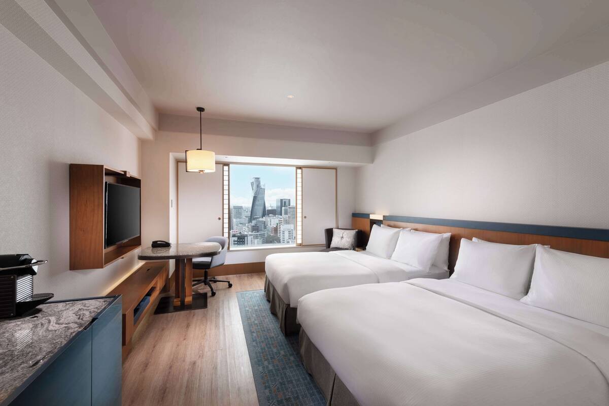 A hotel room with two beds, a desk, a TV, and a city view.