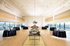 A spacious room with high ceilings and large windows offering a city view. Tables are arranged for a formal event, with a central display and floral arrangements.