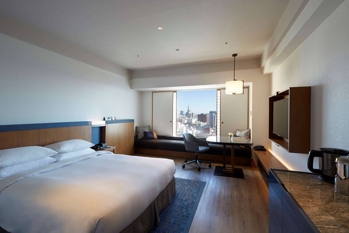A hotel room with a large bed, a desk with a chair, a sofa, a television, and a view of a cityscape.