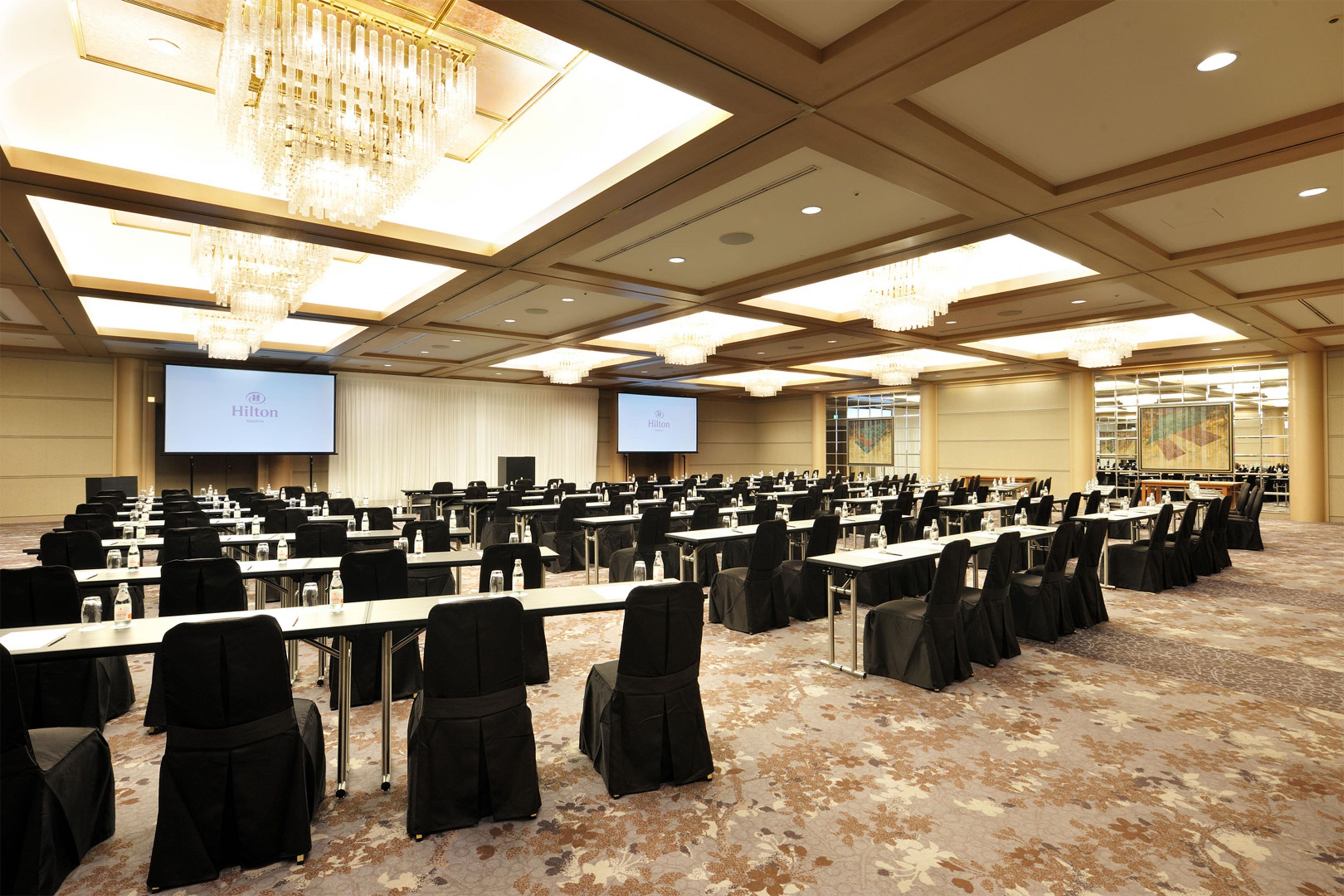 A conference room with rows of tables and chairs, a large screen, and a chandelier.