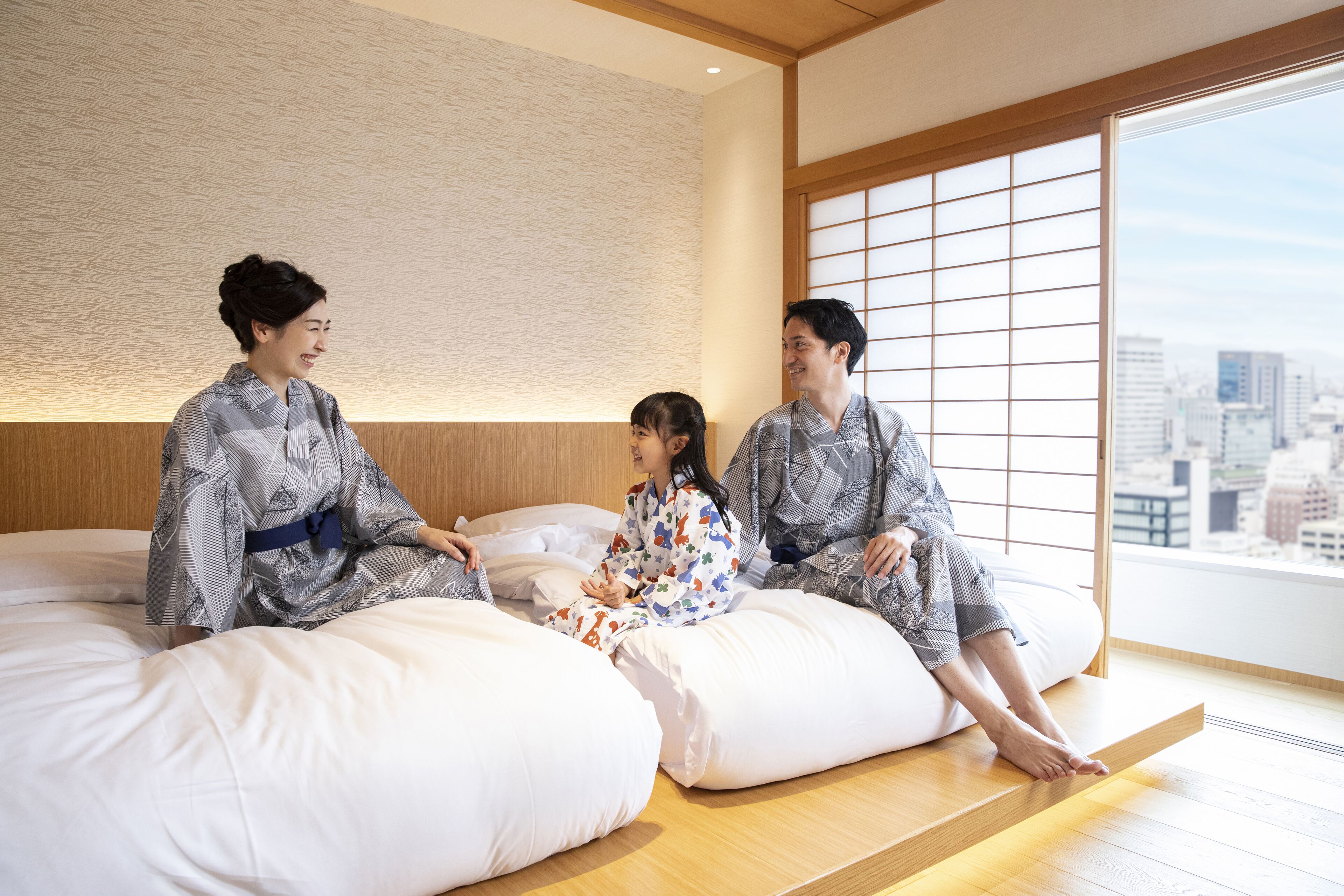 Three people sitting on a low platform bed in a room with a city view.