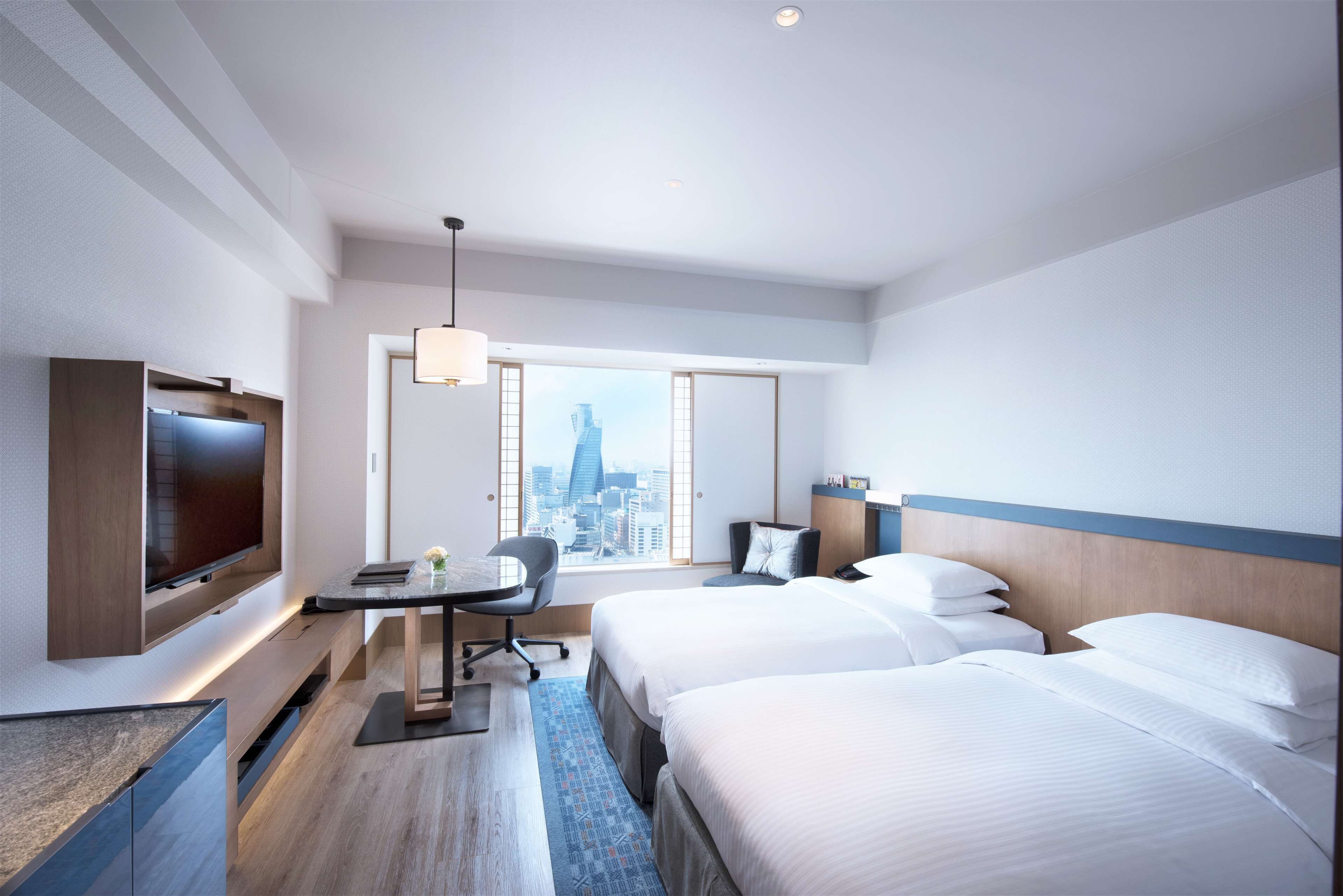 A hotel room with a large TV, a desk with a chair, two beds, a city view, and a modern design.
