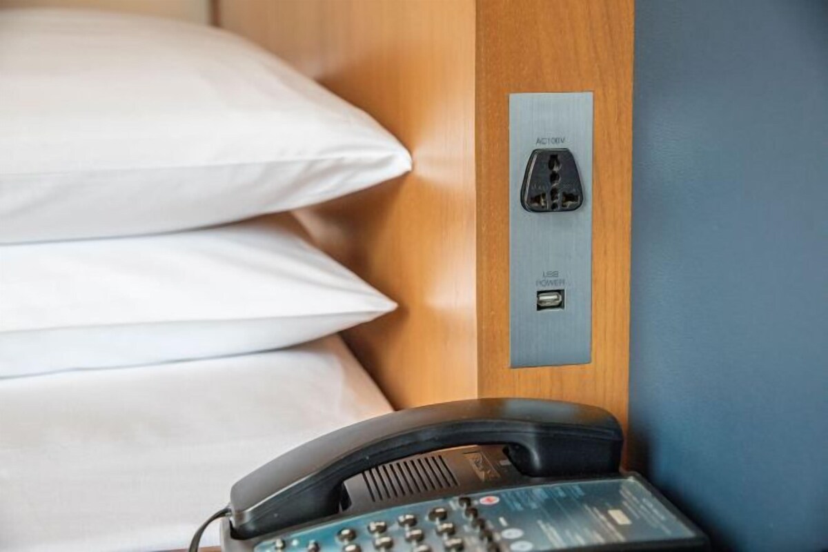 A hotel room with a wooden headboard, a bed with white linens, a telephone, and a wall-mounted USB charging station.