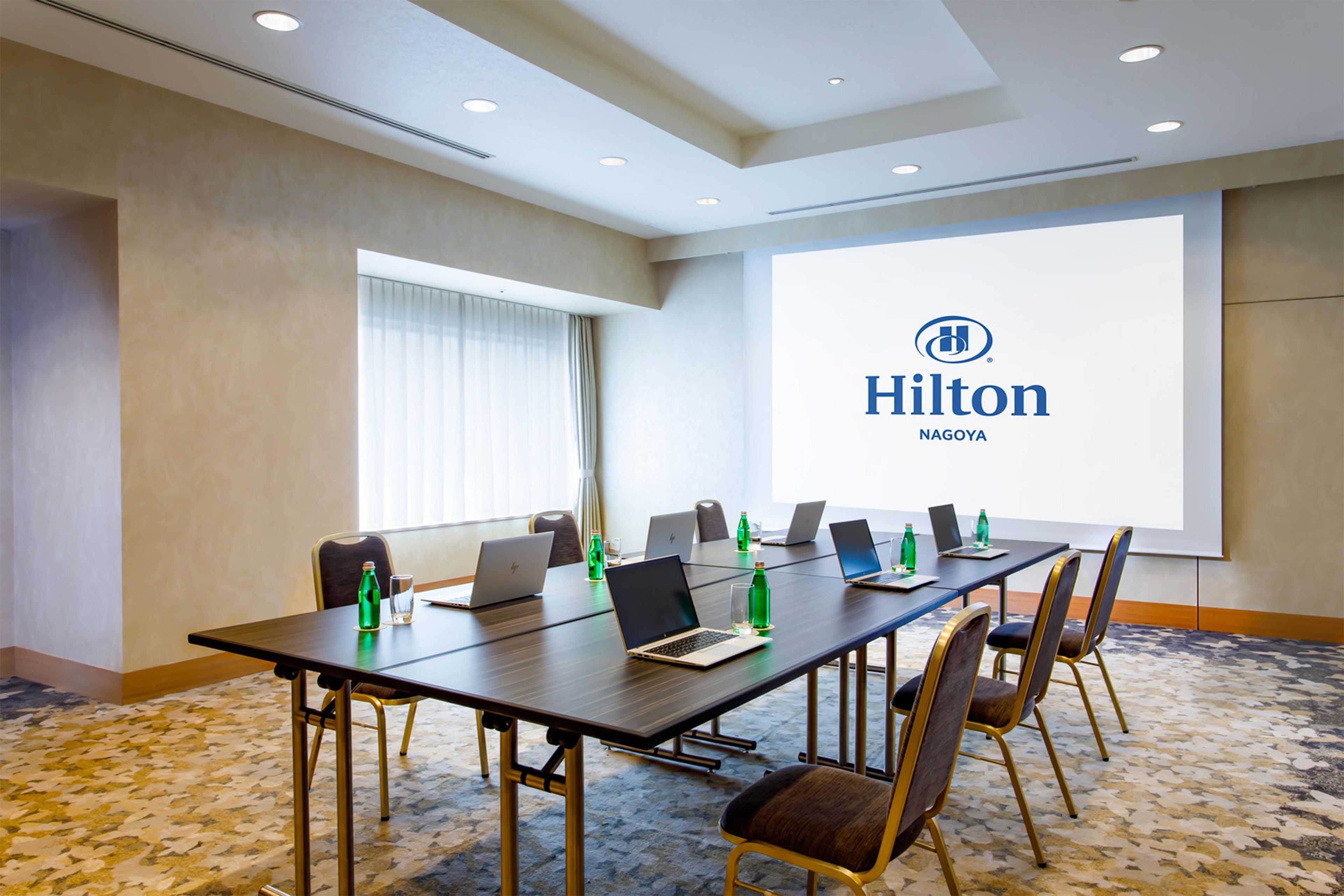Una sala de conferencias con una larga mesa, seis sillas, computadoras portátiles y botellas de agua. Una pantalla proyecta el logotipo de Hilton.