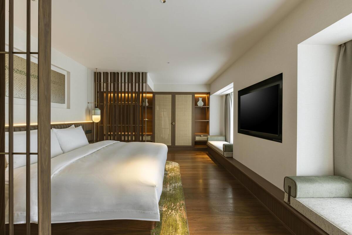A modern hotel room with a large bed, a TV mounted on the wall, and a seating area with a view of the room.