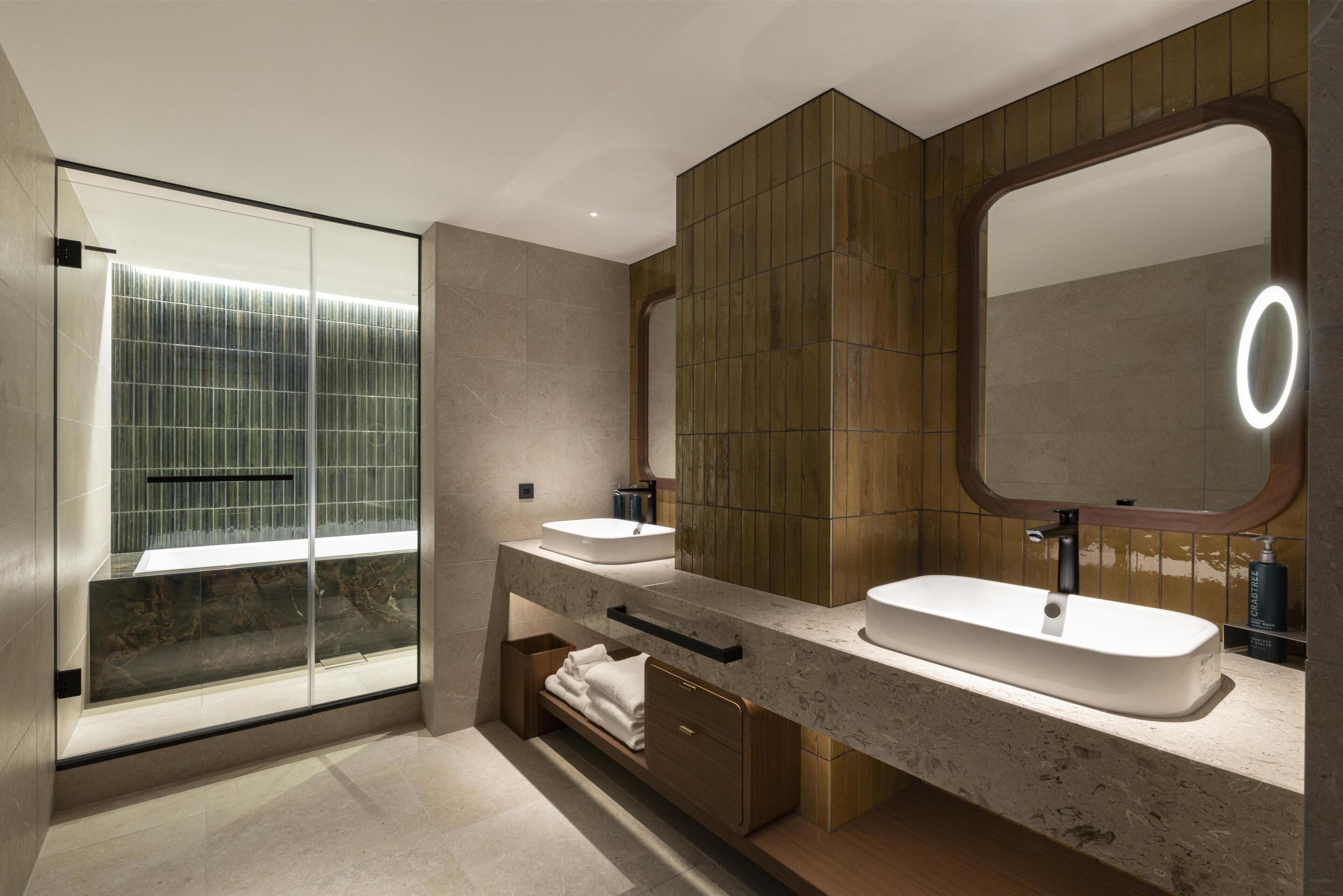 Un baño moderno con cabina de ducha de vidrio, dos lavabos y un espejo grande con luz.