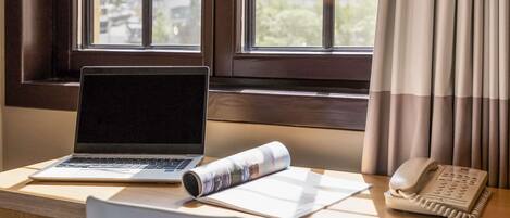 Un escritorio con una computadora portátil, una revista enrollada, un teléfono y una silla frente a una ventana con cortinas.