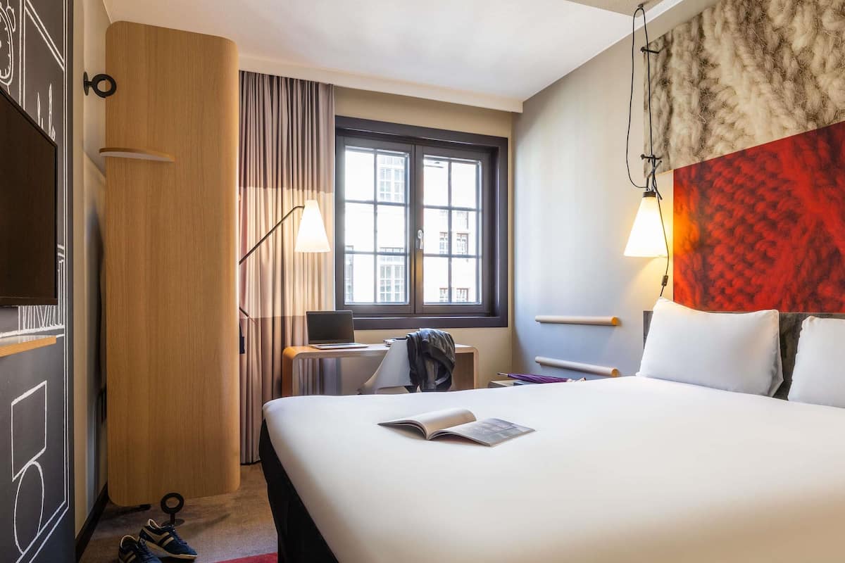 A hotel room with a bed, a desk with a laptop, a window with curtains, a red textured wall, and a pair of shoes on the floor.