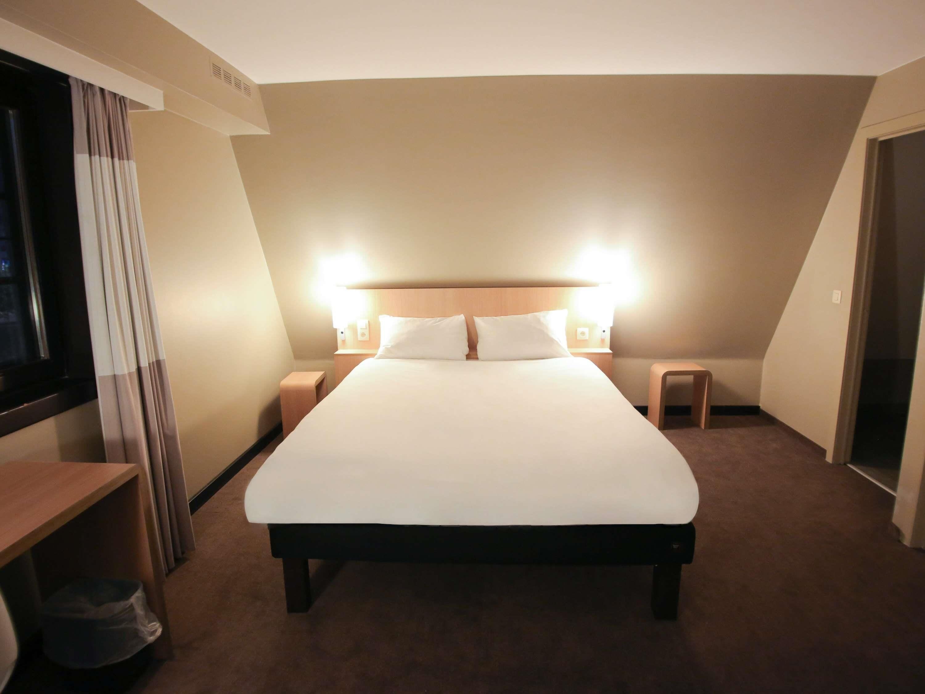 A hotel room with a large bed, two bedside tables, a nightstand, a window with curtains, and a carpeted floor.