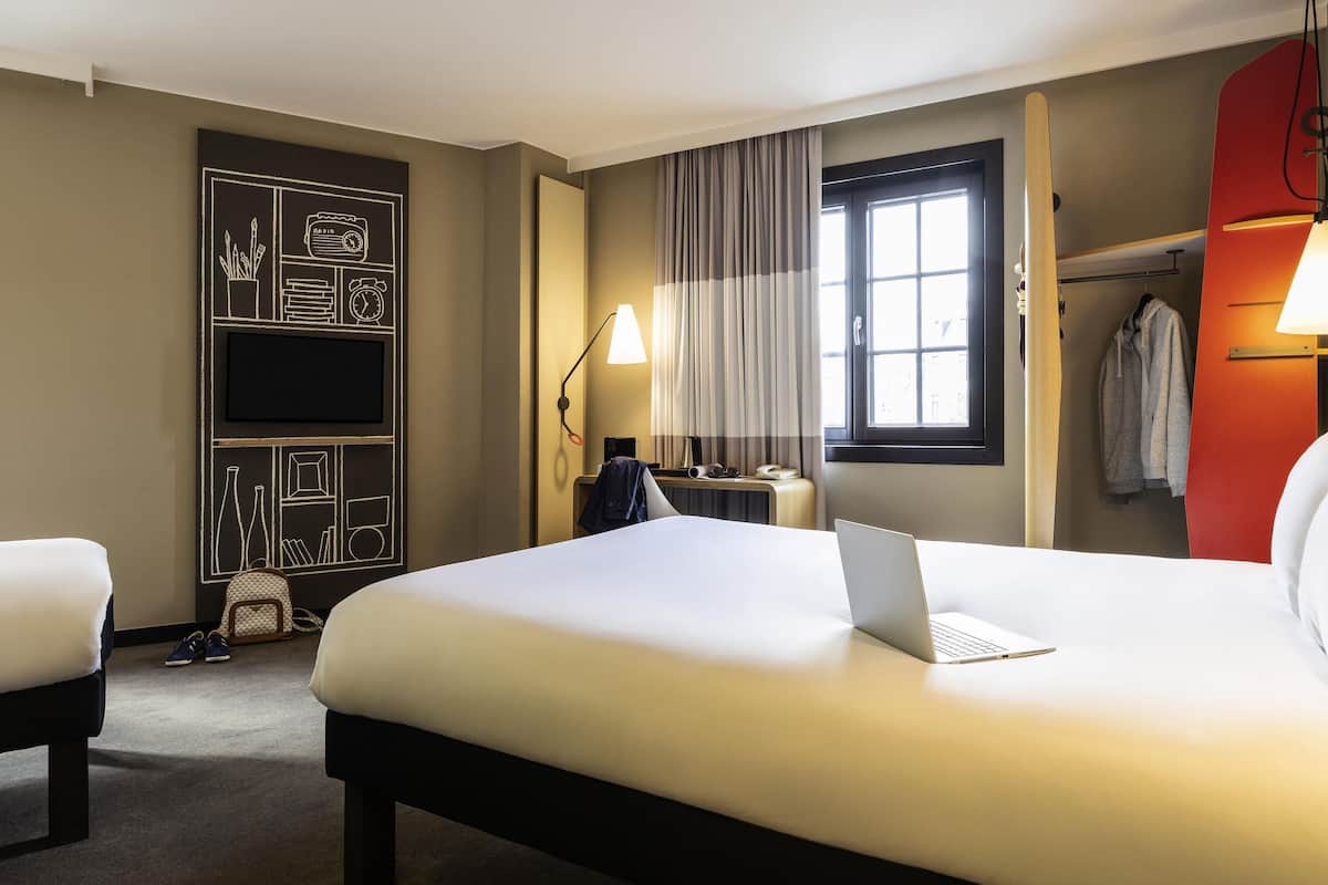 A hotel room with a large bed, a desk with a laptop, a TV, and a window with curtains.