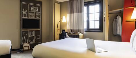 A hotel room with a large bed, a desk with a laptop, a TV, and a window with curtains.