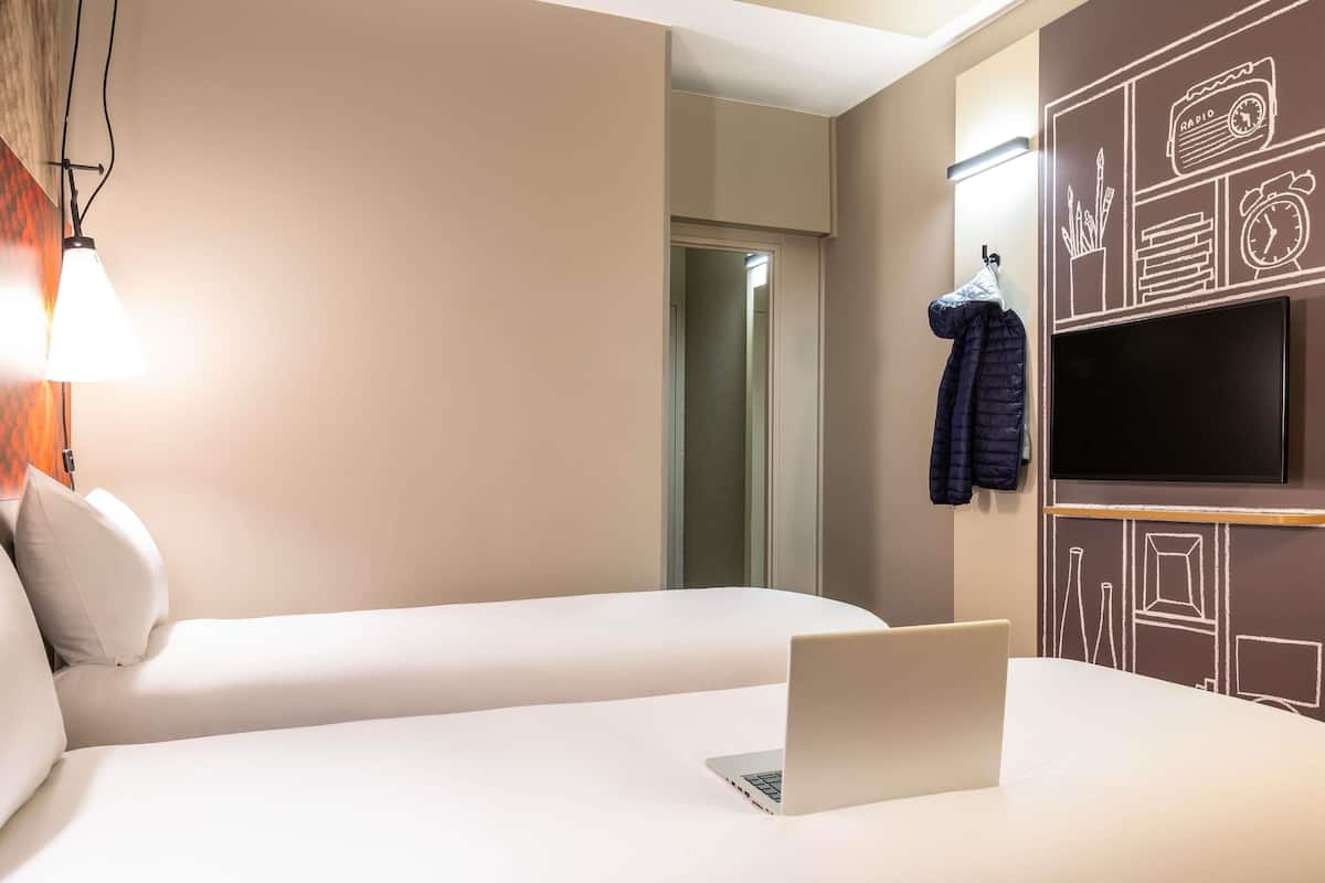 A hotel room with two beds, a laptop on one bed, a TV on the wall, and a coat hanging on the wall.