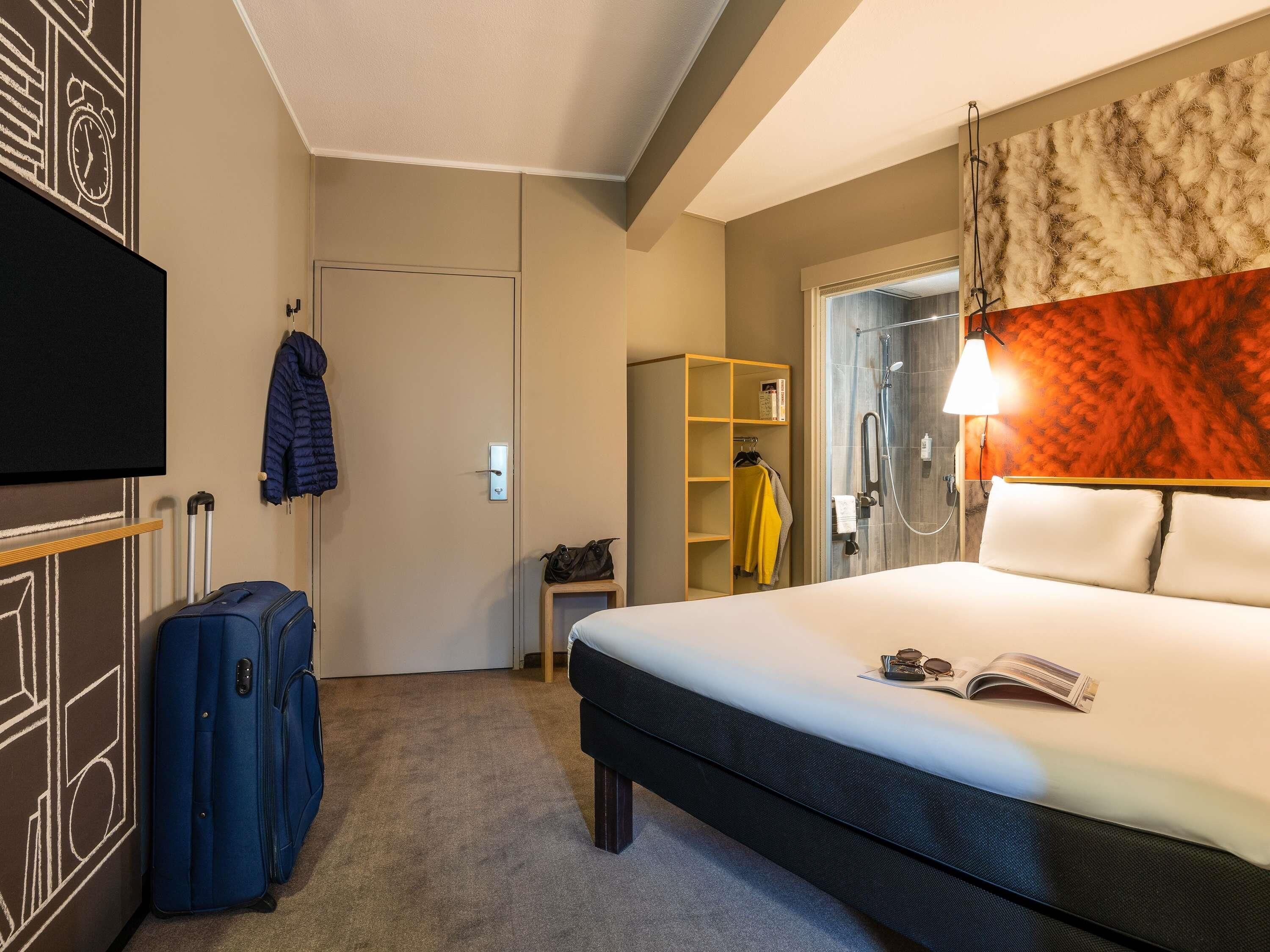 A hotel room with a bed, a suitcase, a coat rack, a wardrobe, and a shower area.