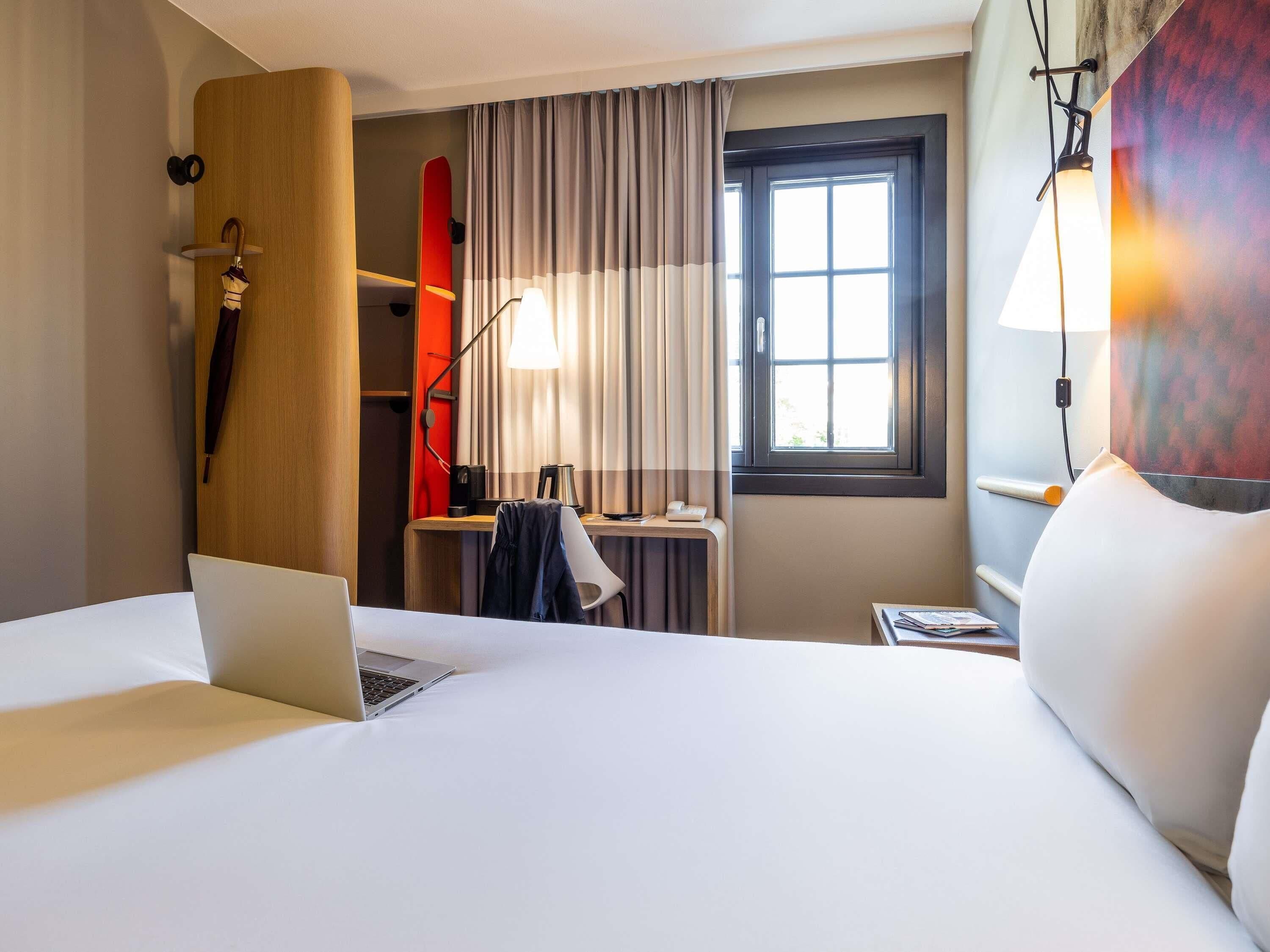 A hotel room with a bed, a desk with a laptop, a surfboard, and a window with curtains.