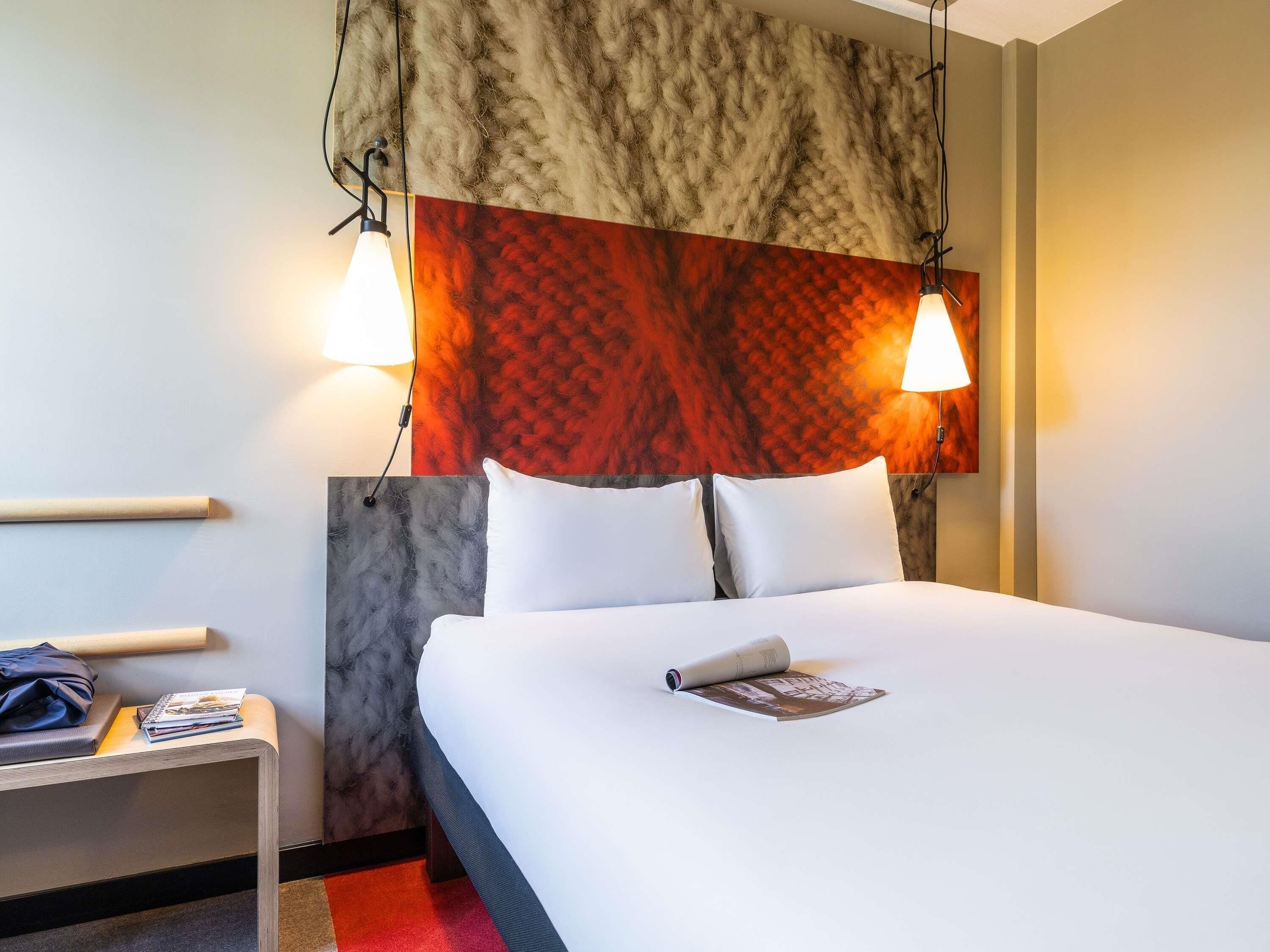 A hotel room with a bed, two bedside lamps, a nightstand with a magazine, and a headboard with a textured pattern.