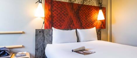 A hotel room with a bed, two bedside lamps, a nightstand with a magazine, and a headboard with a textured pattern.