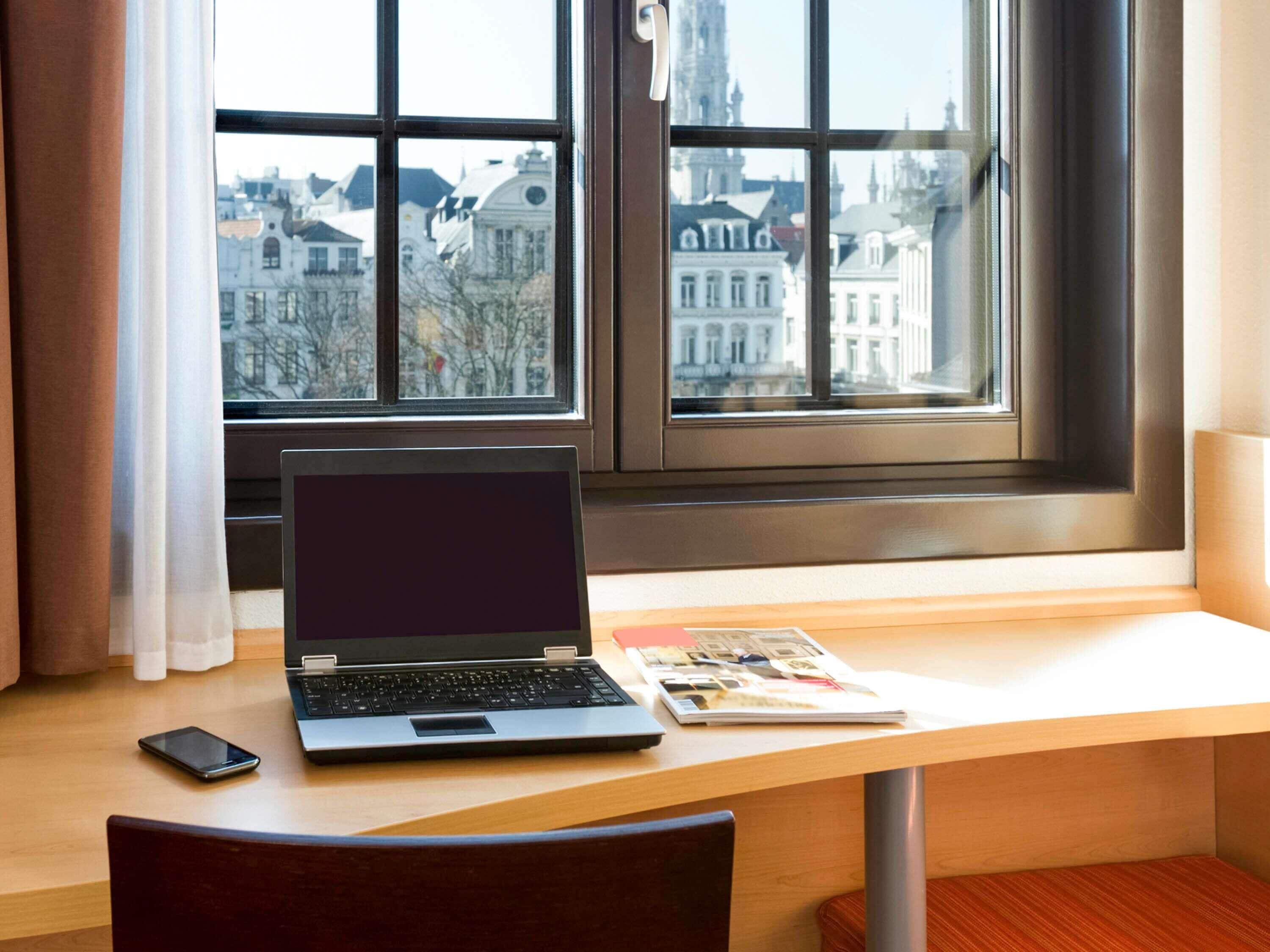 A room with a desk, laptop, and a book. A window offers a view of buildings outside.
