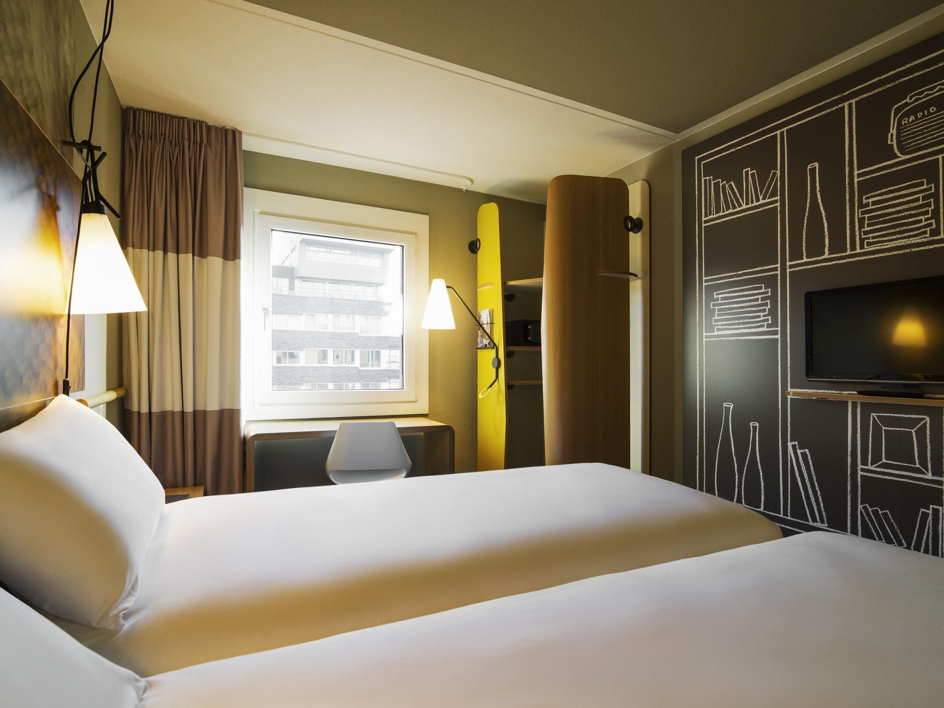 Een hotelkamer met een bed, een bureau, een stoel, een televisie en een raam met uitzicht op gebouwen.