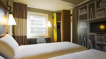 Une chambre d’hôtel équipée d’un lit, d’un bureau, d’une chaise, d’une télévision et d’une fenêtre donnant sur des immeubles.