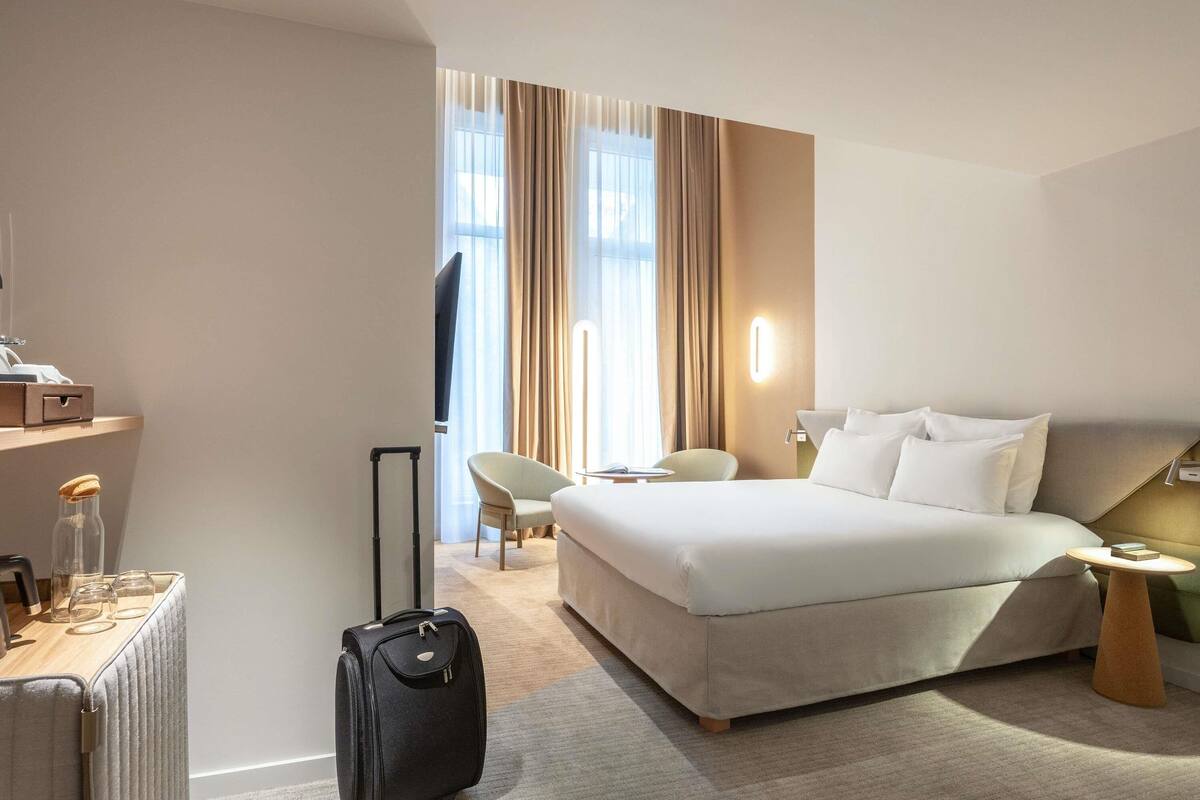 Hotellihuone, jossa on sänky, tuoli, lamppuvarusteinen työpöytä, matkalaukku ja televisio.