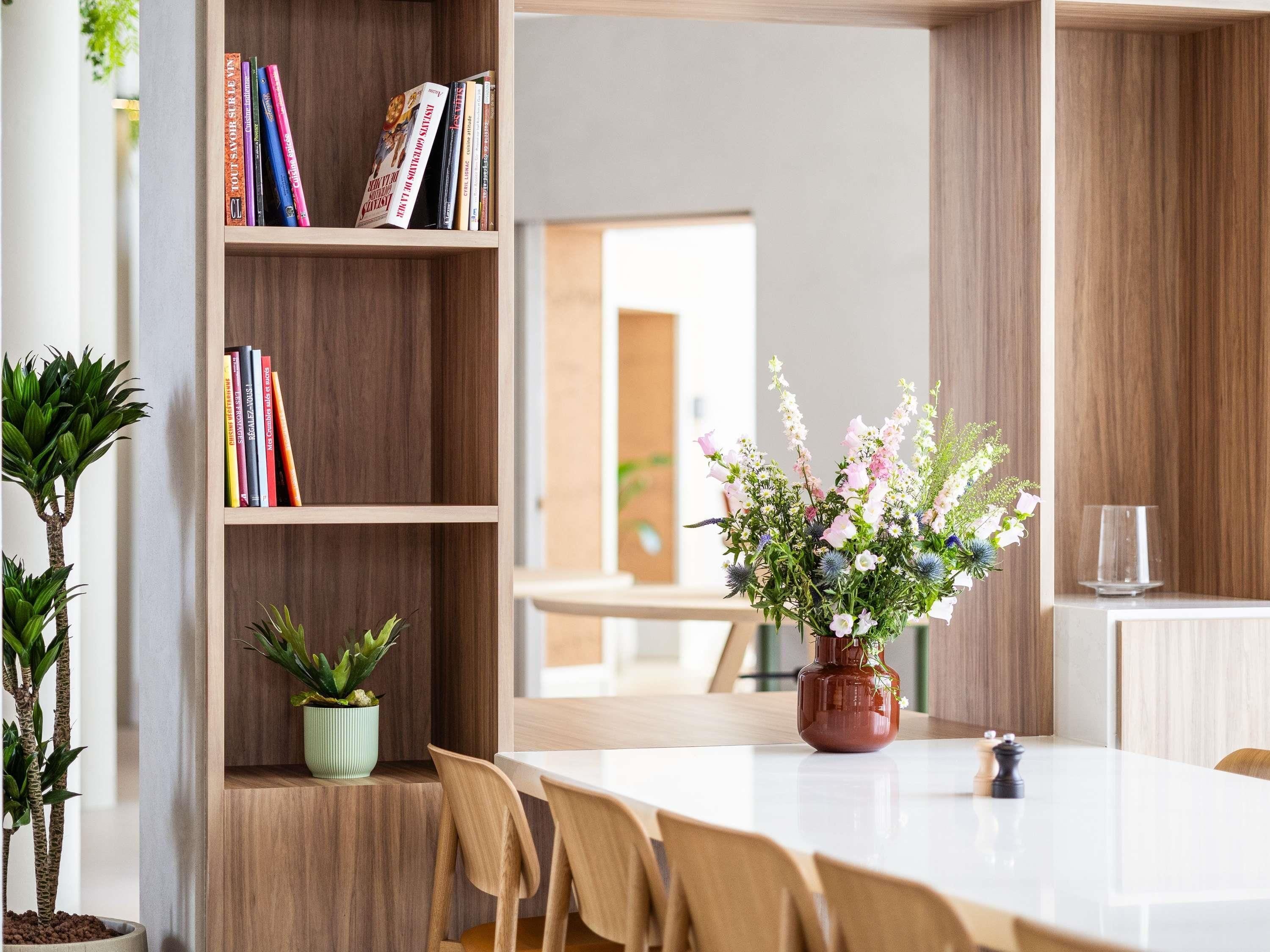 A modern dining area with a wooden bookshelf, a table, chairs, and a vase of flowers.