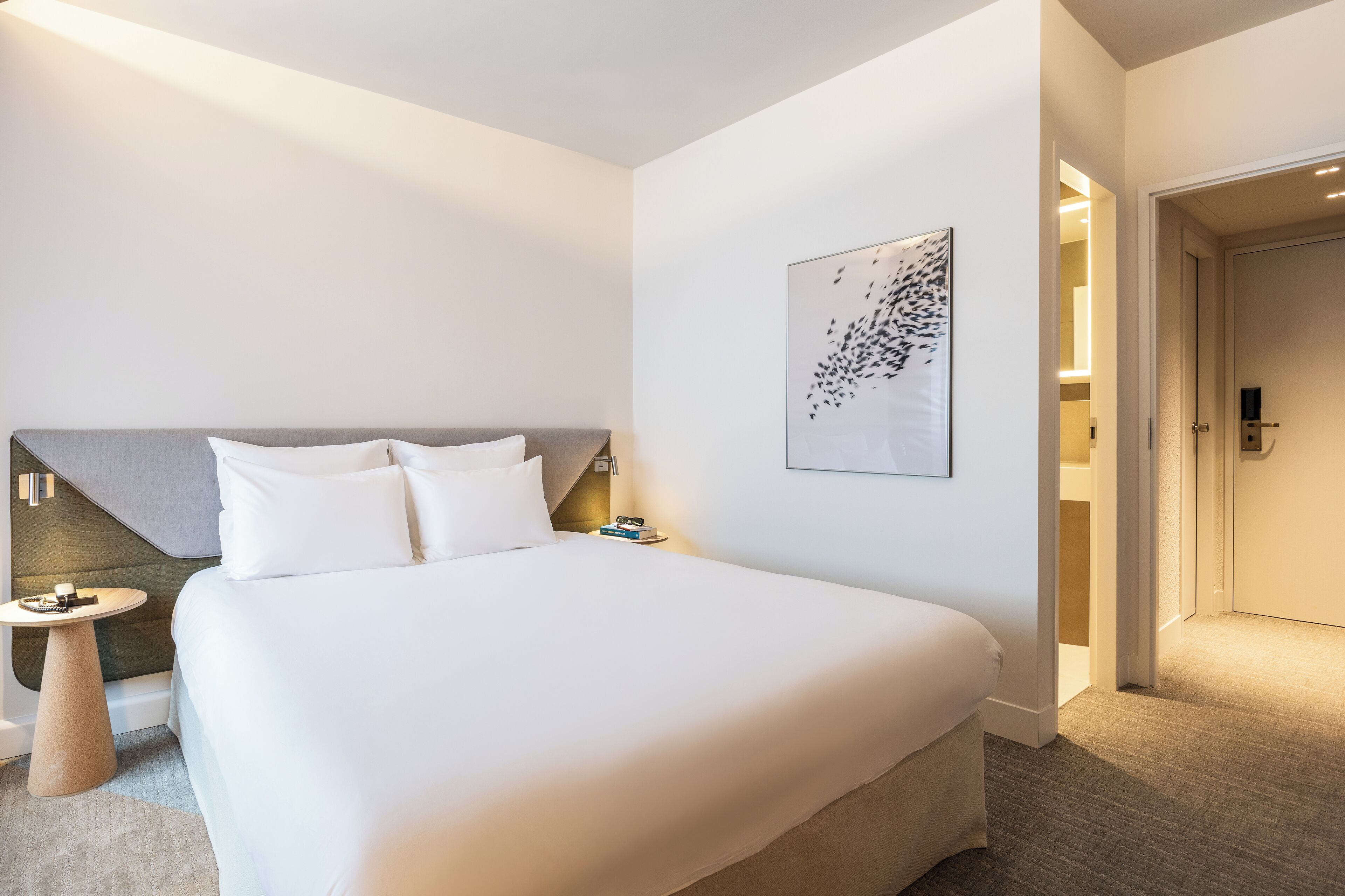 Una camera d'albergo moderna con un letto grande, un comodino, un'opera d'arte astratta appesa al muro e una porta che conduce a un'altra stanza.