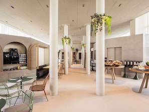 A spacious indoor area with high ceilings, white columns, and hanging plants. There are seating arrangements and a buffet table with food items. - Novotel Brugge Centrum (Bruges)