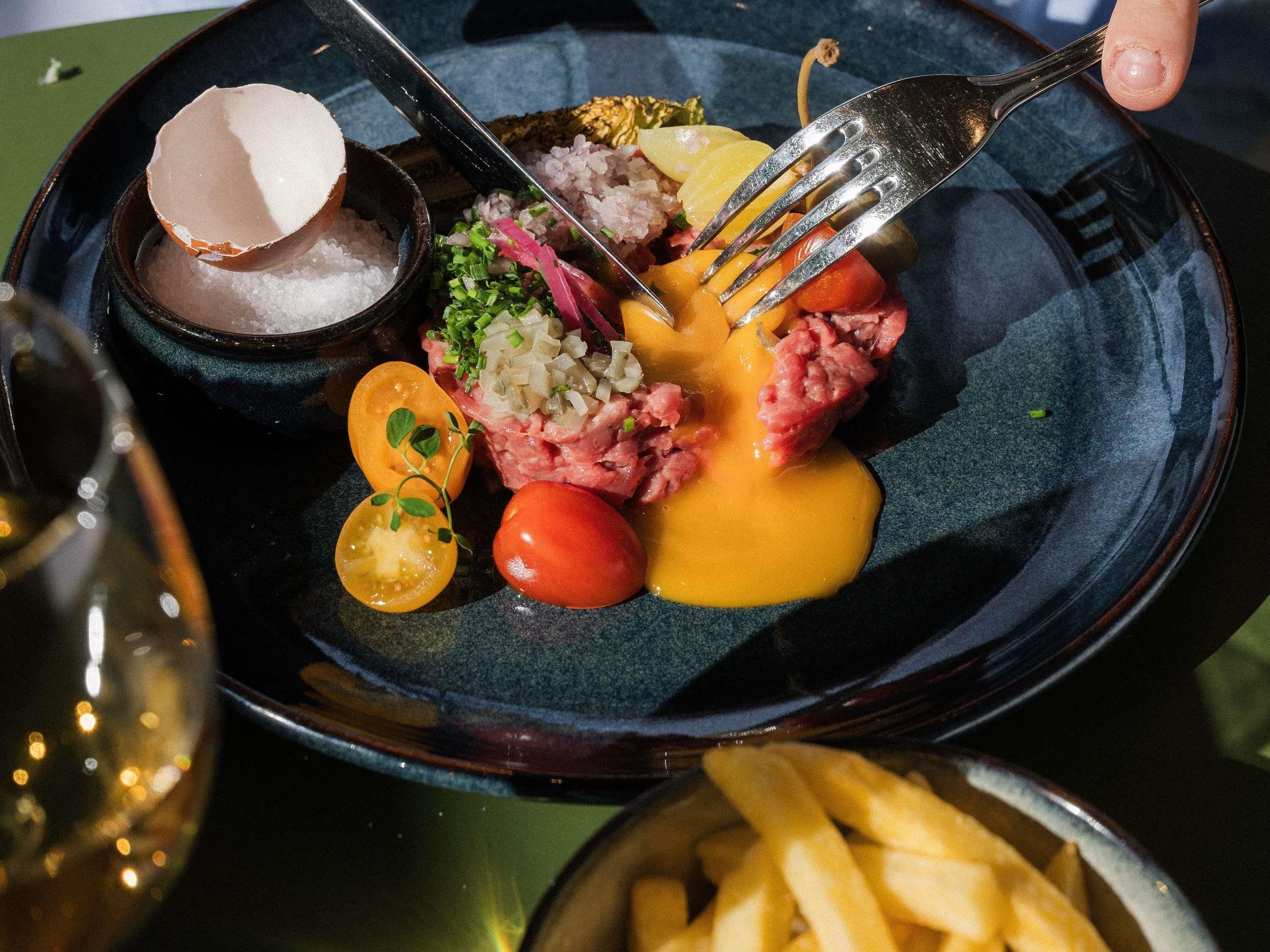 A plate with a steak topped with sauce, garnished with onions and herbs, accompanied by cherry tomatoes and pickled vegetables.
