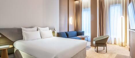 Una habitación de hotel con una cama grande, un sofá, una silla, una mesita y una ventana con cortinas.