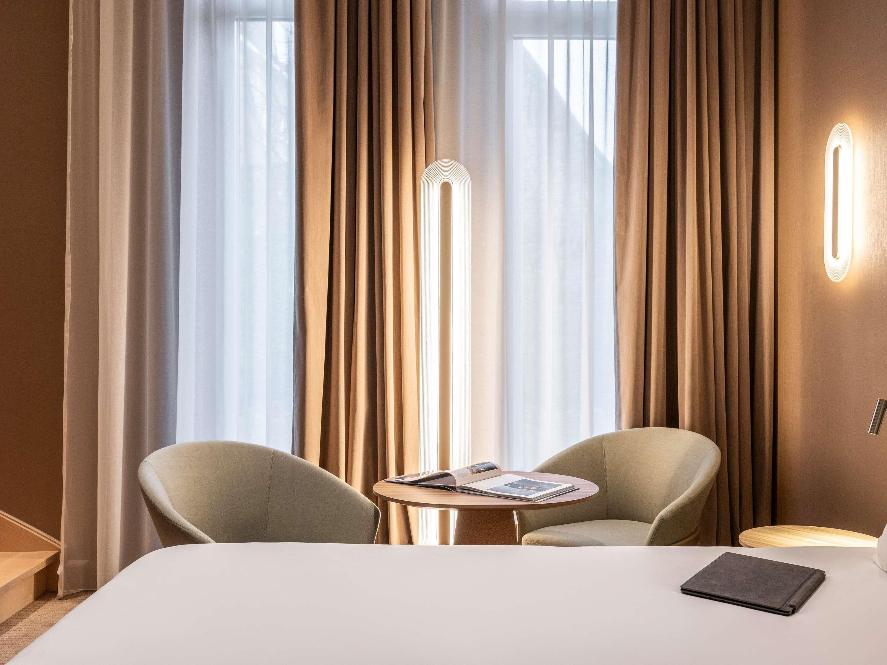 Een hotelkamer met een bed, twee fauteuils, een klein tafeltje met een tijdschrift en een vloerlamp.