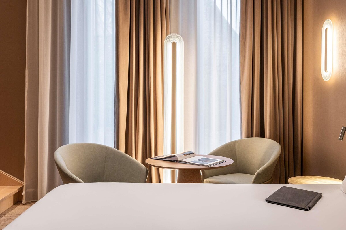 A hotel room with a bed, two armchairs, a small table with a magazine, and a floor lamp.