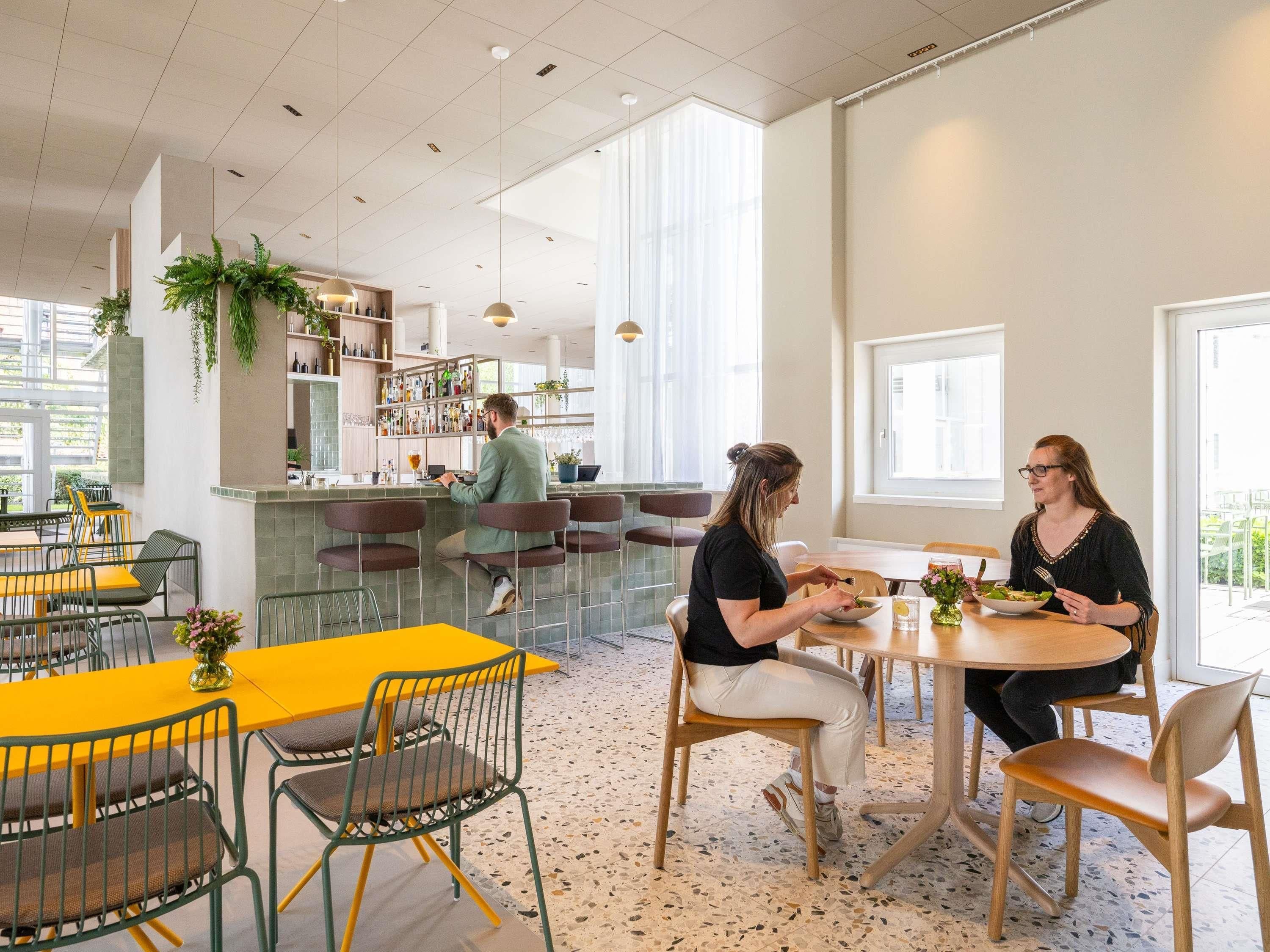 A modern dining area with yellow tables and green chairs, a bar area with shelves of bottles, and large windows allowing natural light.