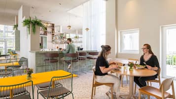 A modern dining area with yellow tables and green chairs, a bar area with shelves of bottles, and large windows allowing natural light.