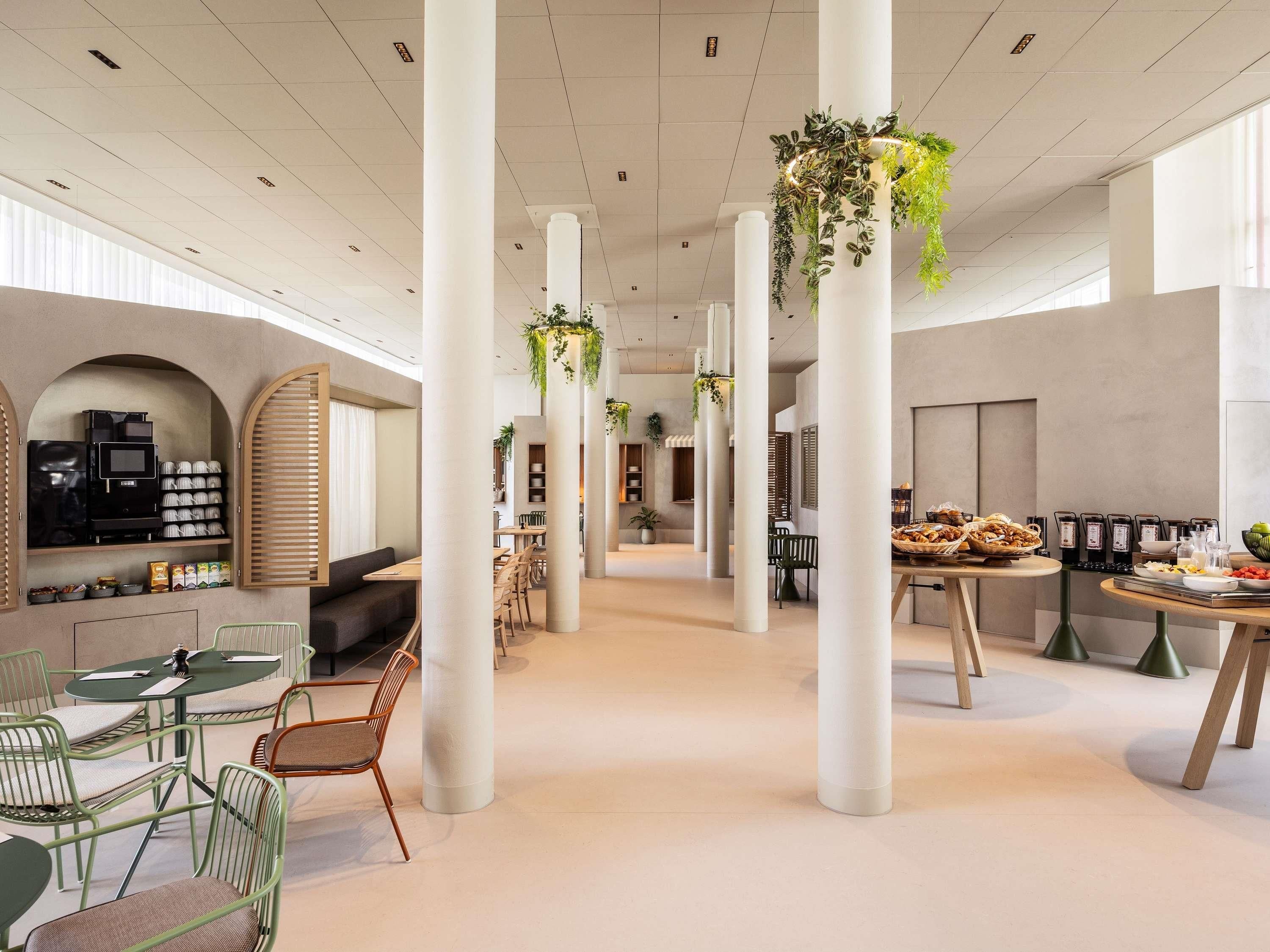 Amplio espacio interior con techos altos, columnas blancas y plantas colgantes. Hay arreglos de asientos y una mesa de buffet con alimentos.