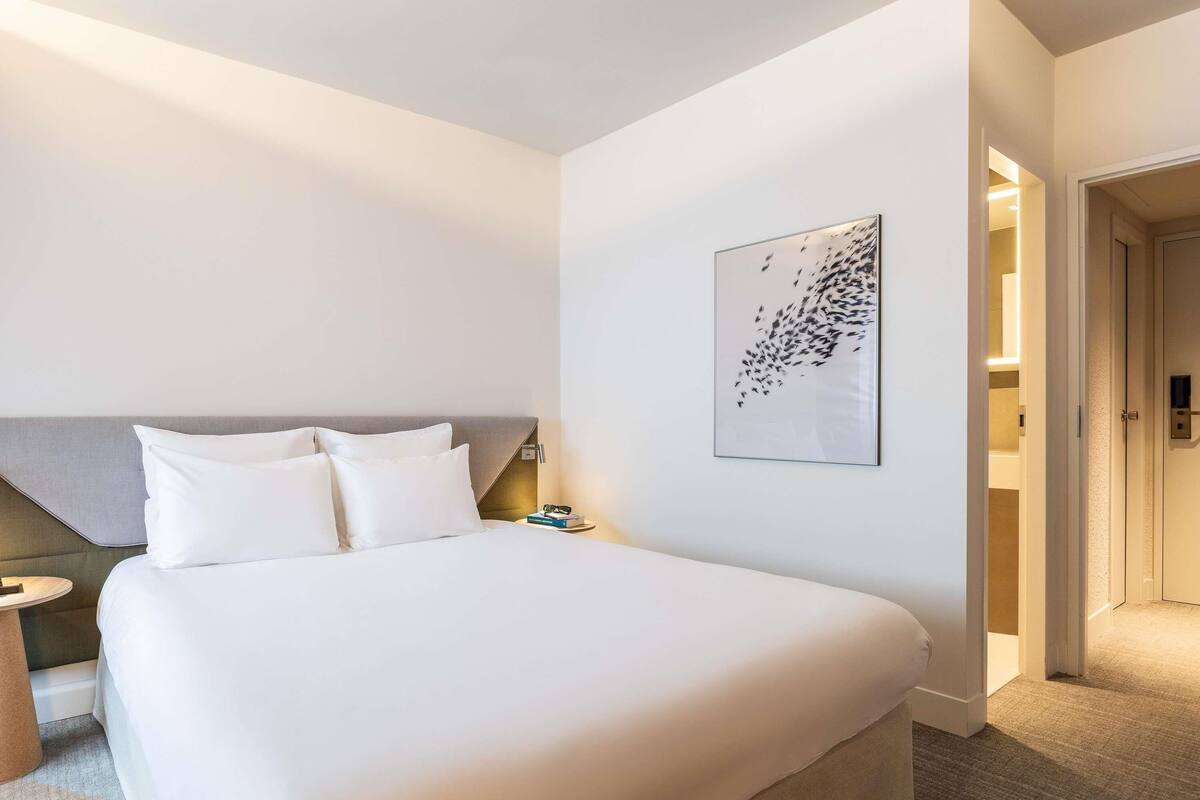 Hotellihuone, jossa on suuri sänky, pääty, yöpöytä, seinälle ripustettu taideteos ja ovi, joka johtaa toiseen huoneeseen.